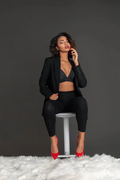 Portrait of a confident black woman with curly dark hair and beautiful makeup sitting by herself on a bed of fur inside a studio with a grey background wearing a black jacket with red high heels.
