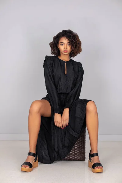 Portrait of a beautiful young black woman with curly hair, beautiful makeup posing by herself sitting indoors wearing a long flowy black dress with black plateau heel sandals and simple jewelry.
