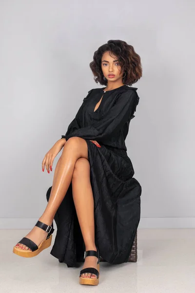 Portrait of a sensual young black woman with curly hair, beautiful makeup posing by herself sitting indoors wearing a long flowy black dress with black plateau heel sandals and simple jewelry.