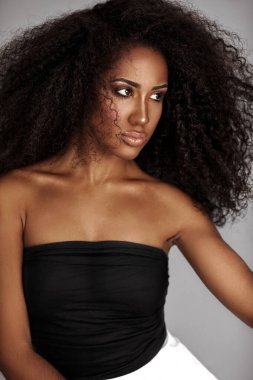 Closeup portrait of a serene young Latin woman with long curly hair and beautiful makeup posing by herself inside a studio with a grey background wearing a black wrapped top and white shorts.