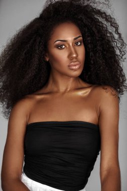 Closeup portrait of a confident young Latin woman with long curly hair and beautiful makeup posing by herself inside a studio with a grey background wearing a black wrapped top and white shorts.