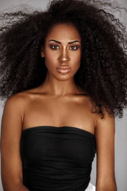 Closeup portrait of a sensual young Latin woman with long curly hair and beautiful makeup posing by herself inside a studio with a grey background wearing a black wrapped top and white shorts.
