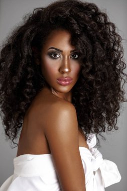 Closeup portrait of a strong young Latin woman with long bouncy curly hair and beautiful makeup posing by herself inside a studio with a grey background wearing only a white drape top.