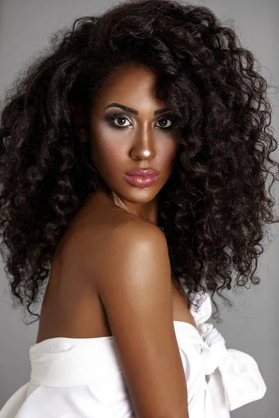 Closeup portrait of a strong young Latin woman with long bouncy curly hair and beautiful makeup posing by herself inside a studio with a grey background wearing only a white drape top.