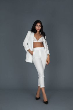 Portrait of a beautiful young white woman with wavy black hair and beautiful makeup posing by herself inside a studio with a grey background wearing a white suit with black high heels and a white bra.