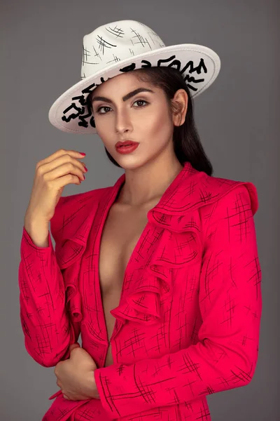 Closeup portrait of a beautiful young white woman with wavy black hair and beautiful makeup posing by herself inside a studio with a grey background wearing a red business suit with a white hat.