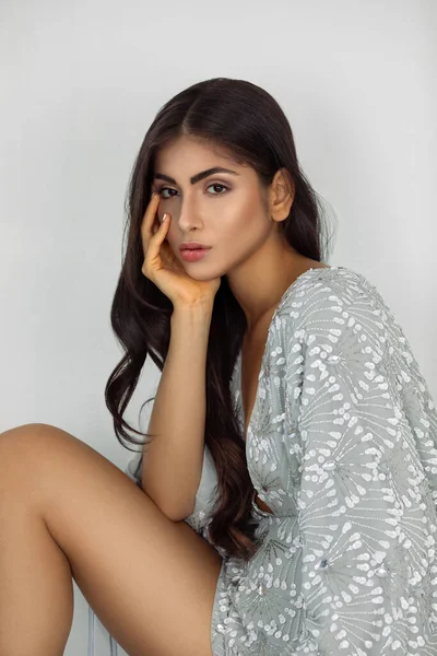 Closeup portrait of a beautiful young white woman with wavy black hair and beautiful makeup sitting by herself inside a studio with a white background wearing a short silver dress with high heels.
