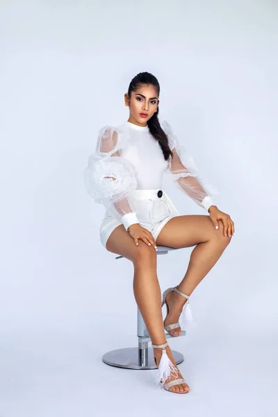 Portrait of a sexy young white woman with black hair and beautiful makeup sitting by herself inside a studio with a white background wearing white shorts with a flowy blouse and high heels.