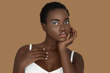 Closeup portrait of a sexy young black woman with short Afro hair, light makeup and lipstick posing by herself resting her head on the palm of her hand inside a studio with a pecan background.