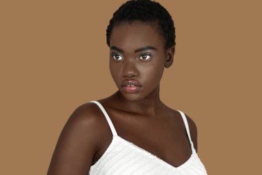 Closeup portrait of a serene young black woman with short Afro hair, light makeup and lipstick posing by herself inside a studio with a pecan background wearing a white strapped dress.