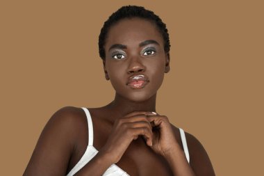 Closeup portrait of a cute young black woman with short Afro hair, light makeup and lipstick posing by herself inside a studio with a pecan background wearing a white strapped dress.