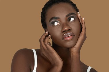 Closeup portrait of a cute young black woman with short Afro hair, light makeup and lipstick posing by herself inside a studio with a pecan background resting the palms of her hands on her face.