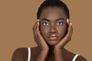 Closeup portrait of a sensual young black woman with short Afro hair, light makeup and lipstick posing by herself inside a studio with a pecan background resting the palms of her hands on her face.