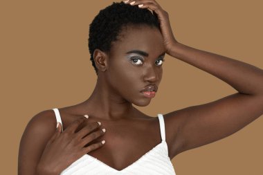 Closeup portrait of a sensual young black woman with short Afro hair, light makeup and lipstick posing by herself inside a studio with a pecan background resting the palm of her hand on her head.