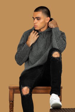 Portrait of a serene young Latin man with short hair, light makeup and eyeshadow sitting by himself inside a studio with a pecan background wearing black jeans and a grey hooded sweater.
