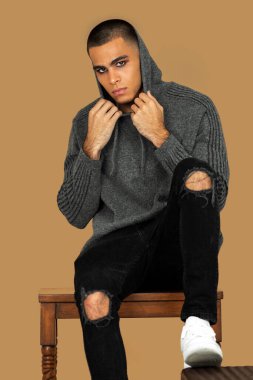 Portrait of a confident young Latin man with short hair, light makeup and eyeshadow sitting by himself inside a studio with a pecan background wearing black jeans and a grey hooded sweater.