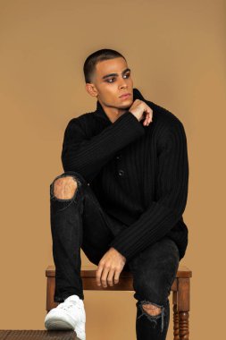 Portrait of a thoughtful young Latin man with short hair, light makeup and eyeshadow sitting by himself inside a studio with a pecan background wearing black jeans and a black sweater.