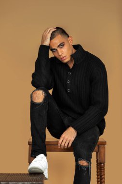 Portrait of a strong young Latin man with short hair, light makeup and eyeshadow sitting by himself inside a studio with a pecan background wearing black jeans and a black sweater.
