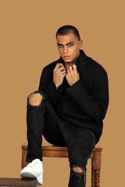 Portrait of a confident young Latin man with short hair, light makeup and eyeshadow sitting by himself inside a studio with a pecan background wearing black jeans and a black sweater.
