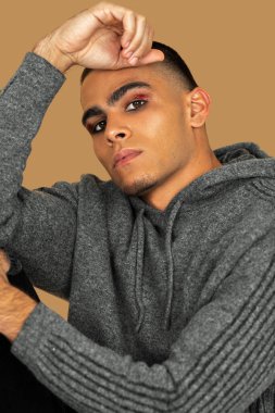 Closeup portrait of an attractive Latin man with short hair, light makeup and eyeshadow sitting by himself inside a studio with a pecan background wearing black jeans and a grey hooded sweater.
