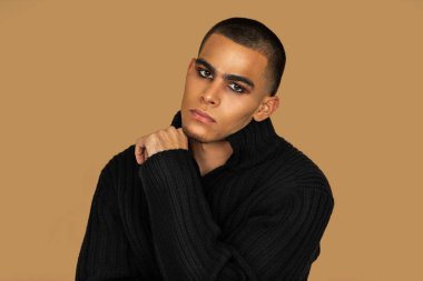 Closeup portrait of a handsome young Latin man with short hair, light makeup and eyeshadow sitting by himself inside a studio with a pecan background wearing black jeans and a black sweater.