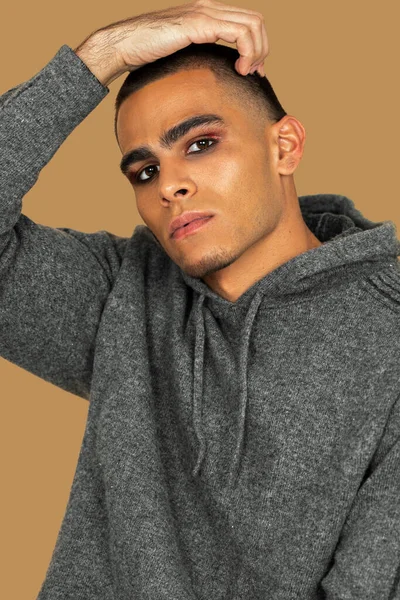 Portrait of a sexy young Latin man with short hair, light makeup and eyeshadow sitting by himself inside a studio with a pecan background wearing black jeans and a grey hooded sweater.