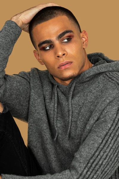 Closeup portrait of a young Latin man with short hair, light makeup and eyeshadow sitting by himself inside a studio with a pecan background wearing black jeans and a grey hooded sweater.