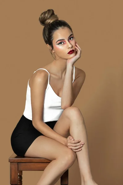 Portrait of a beautiful young Latina with long dark blond hair, eye makeup and lipstick sitting by herself on a table inside a studio with a pecan background wearing a white strap top and black skirt.
