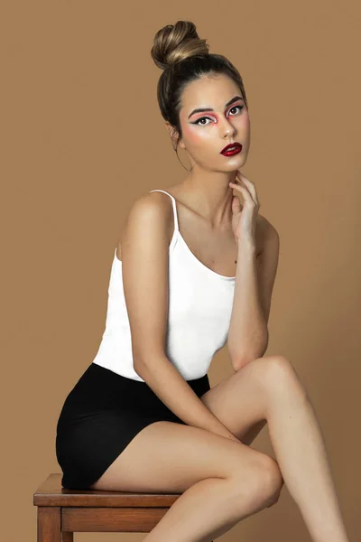 Portrait of a sensual young Latina with long dark blond hair, eye makeup and lipstick sitting by herself on a table inside a studio with a pecan background wearing a white strap top and black skirt.
