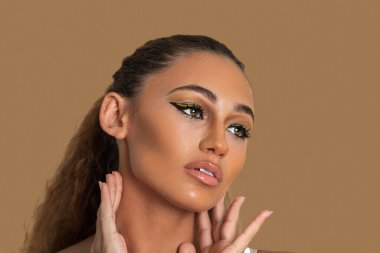 Closeup portrait of a lovely young mixed race woman with long blond hair, light makeup and eye shadow posing by herself touching her head inside a studio with a pecan background.