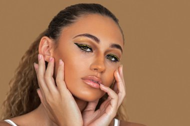 Closeup portrait of a young thoughtful mixed race woman with long blond hair, light makeup and eye shadow posing by herself touching her head and lips inside a studio with a pecan background.