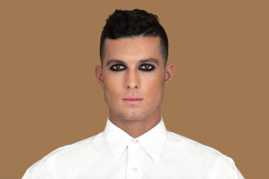 Closeup portrait of a handsome young White man with short brown hair, light makeup and dark eyeshadow sitting by himself inside a studio with a pecan background wearing a white button up shirt.