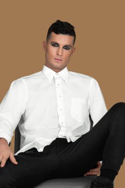 Portrait of a young White man with short brown hair, light makeup and dark eyeshadow sitting by himself inside a studio with a pecan background wearing a white button up shirt and black pants.
