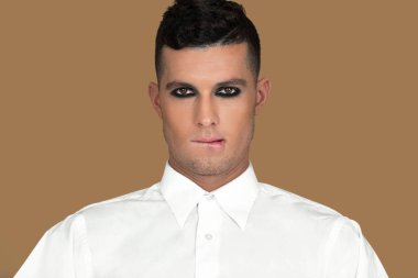 Closeup portrait of a playful young White man with short brown hair, light makeup and dark eyeshadow sitting by himself inside a studio with a pecan background wearing a white button up shirt.