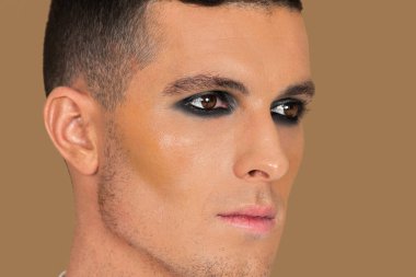 Closeup side portrait of a young White man with short brown hair, light makeup and dark eyeshadow sitting by himself inside a studio with a pecan background wearing a white button up shirt.