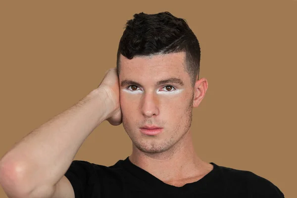 Portrait of a handsome young White man with short brown hair, light makeup and white eyeshadow sitting by himself inside a studio with a pecan background wearing a black shirt.