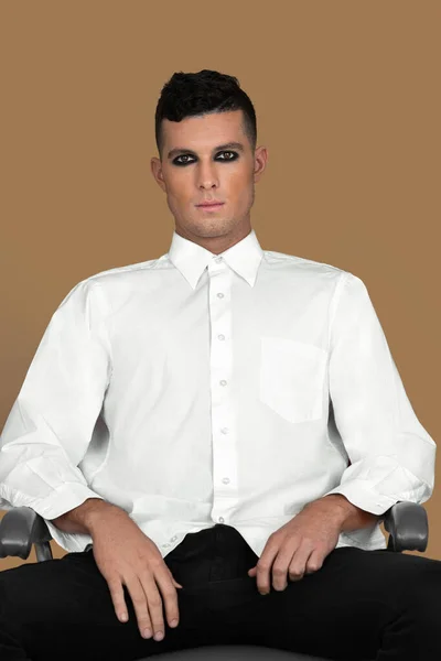 Portrait of a sexy young White man with short brown hair, light makeup and dark eyeshadow sitting by himself inside a studio with a pecan background wearing a white button up shirt and black pants.