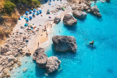 Mavi denizin havadan görünüşü, güneşli yaz günlerinde şemsiyeli kumlu plaj, kayalar ve kayalar. Lefkada Adası, Yunanistan. Deniz kıyılı tropik bir manzara, yüzen insanlar, mavi su. Üst görünüm