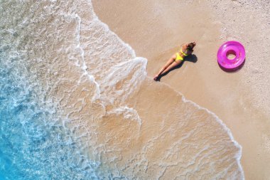 Gün batımında dalgalarla birlikte kumlu sahilde pembe yüzme yüzüğüyle uzanan genç ve güzel kadının havadan görüntüsü. Yunanistan 'ın Lefkada adasında yaz tatili. Seksi kız manzarası, berrak mavi su.