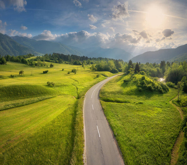 Aerial view of road in green meadows and hils at sunset in summer. Top view from drone of rural road, mountains, forest. Beautiful landscape with roadway, trees, green grass, sky with clouds. Slovenia