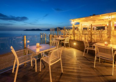 Beautiful cafe on sea coast at night in summer. Landscape with chairs and tables, lamps, lights, water, blue sky at dusk. Empty cozy cafe outdoor with illumination in Oludeniz, Turkey. Travel