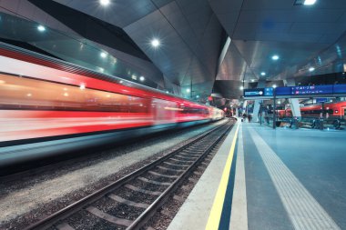 High speed train in motion on the railway station at night. Moving red modern intercity passenger train, railway platform, architecture, city lights. Modern train station in Vienna, Austria. Railroad