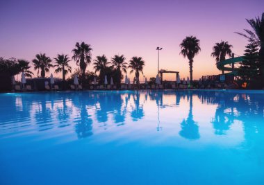 Beautiful reflection in swimming pool at colorful sunset. Purple sky reflected in water, palm trees, sun beds, umbrellas at night in summer. Luxury resort. Landscape with empty pool in twilight