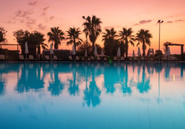 Beautiful reflection in swimming pool at colorful sunset. Purple sky reflected in water, palm trees, sun beds, umbrellas at night in summer. Luxury resort. Landscape with empty pool in twilight