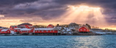 Norwegian fishing village and sea coast at sunset. Lofoten islands, Norway. Winter landscape with red houses, blue water, dramatic cloudy sky with golden sun beams. Norwegian traditional red rorbu