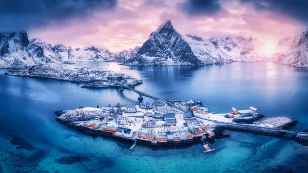 Aerial view of snowy islands with houses, rorbu, blue sea, mountains, bridge and ponk cloudy sky at sunset in winter. Dramatic landscape with village, rock, road. Top view. Lofoten islands, Norway