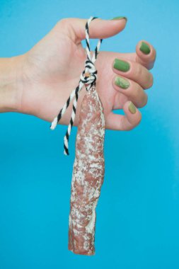 Sosis fuet kadınların parmaklarında asılı. Kız sosisi ipte tutuyor. yakın plan ve mavi arkaplan