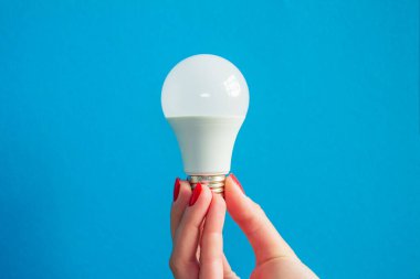 Bir kadının elindeki lamba. LED lamba tutan kadın parmakları. Bir fikrim var. Beyaz lamba. Kırmızı manikür. mavi arka plan. Yatay resim.