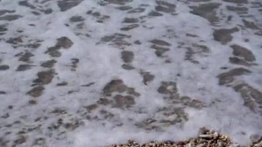 Beyaz çakıl taşlı bir plajın arka planında köpükle denizin şeffaf dalgaları.