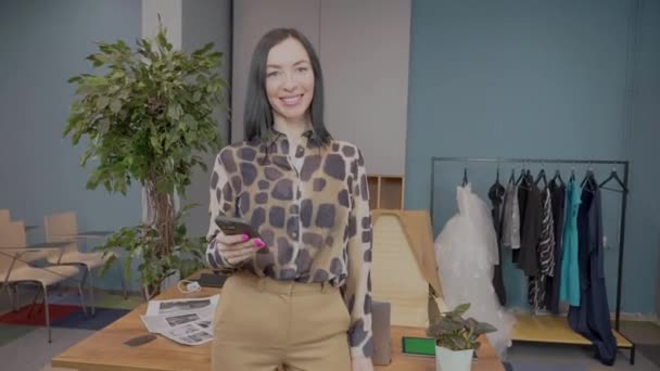 Portrét krásné usmívající se evropské módní návrhářky stojící v ateliéru, pózující pro kameru s telefonem. Žena krejčí hrdý a spokojenější se svou prací. — Stock video